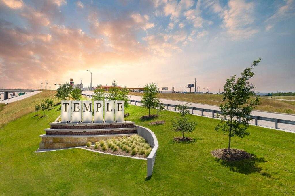 Entrance sign for Temple, Texas along a landscaped roadside with trees, highway access, and a sunset sky in the background.