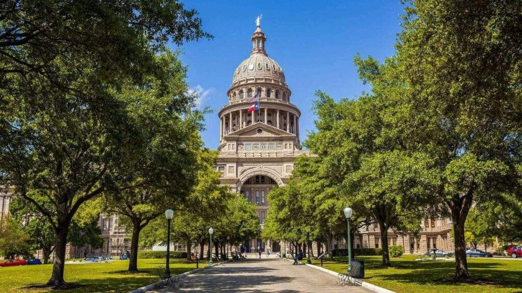 Texas State Capitol building in Austin, Texas, framed by tree-lined walkways, featuring its iconic dome and historic architecture under a clear blue sky.