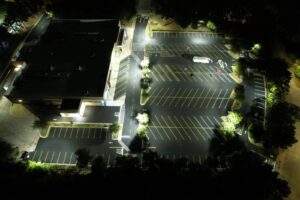 Aerial nighttime view of a newly paved commercial asphalt parking lot with fresh striping, directional markings, and bright LED lighting surrounding a large building.