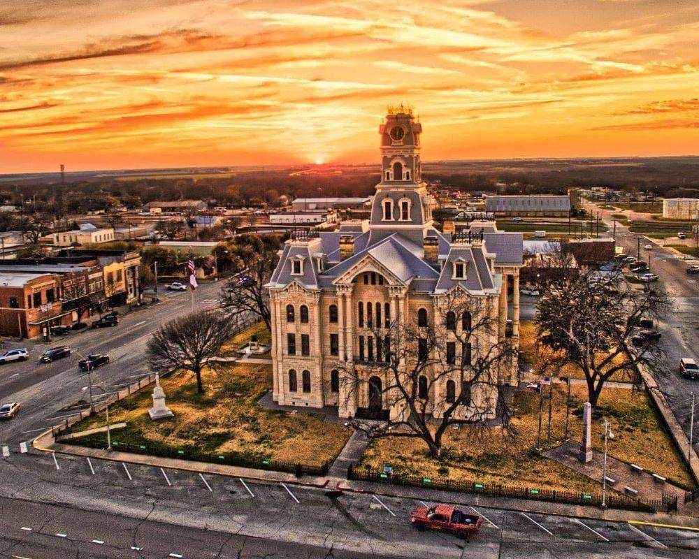 Historic courthouse in downtown Gatesville, Texas, photographed from above at sunset, featuring classic limestone architecture, a prominent clock tower, surrounding streets, and a vibrant orange sky over the community.
