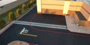 Freshly paved commercial parking lot with newly painted pavement markings, crosswalk striping, fire lane boundaries, and directional traffic lines surrounding a retail building, showcasing a clean and well-organized parking area.
